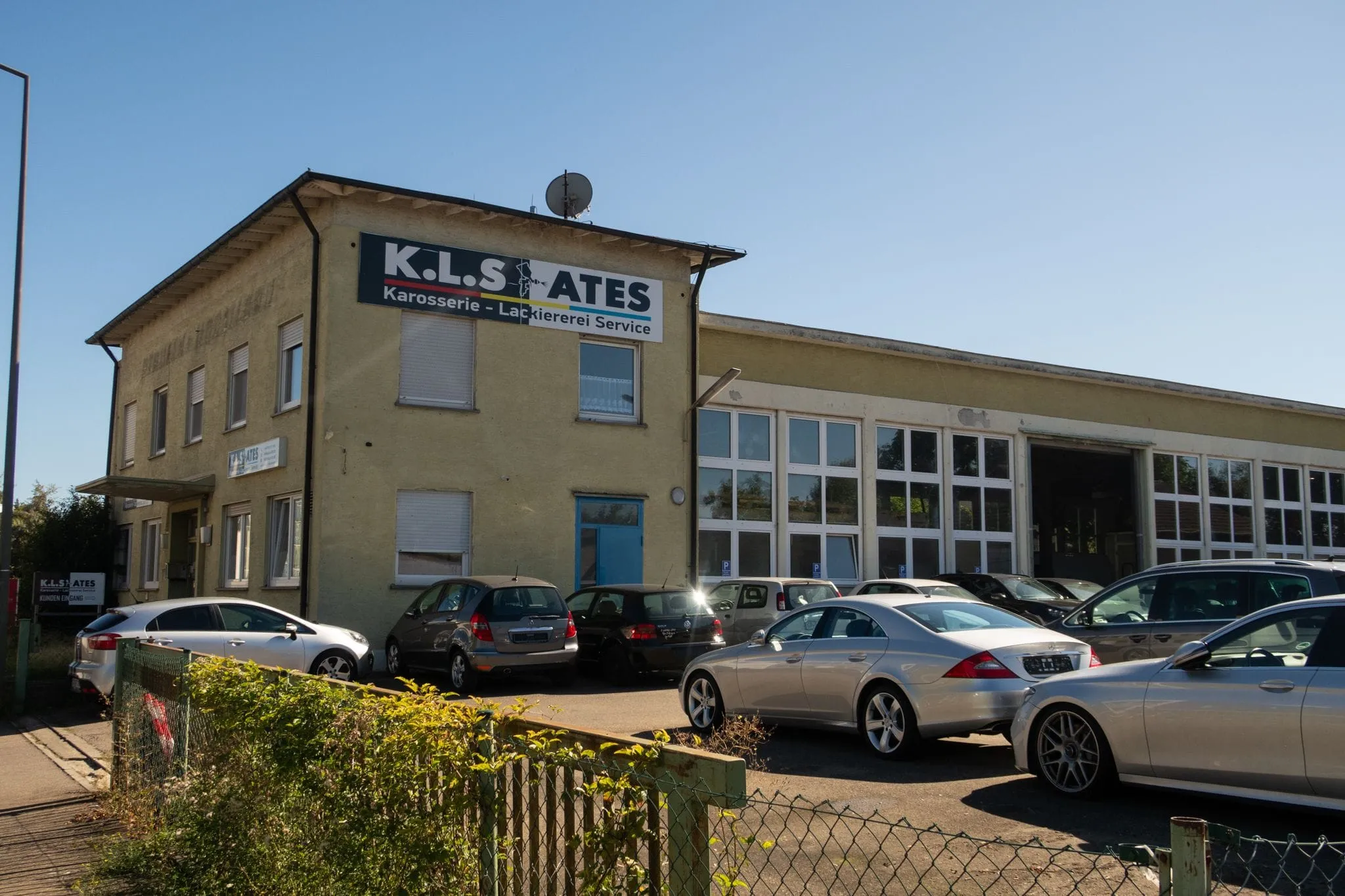 Firmengebäude von K.L.S. Ates mit großem Firmenschild an der Fassade, rechts die Werkstatthalle mit Fensterfront und Einfahrt, davor Kundenfahrzeuge